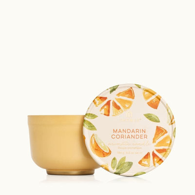 The Mandarin Coriander Poured Candle Tin by Thymes features a yellow container and lid adorned with watercolor illustrations of orange slices and leaves, labeled Mandarin Coriander Aromatic Candle, offering an enchanting burn time.