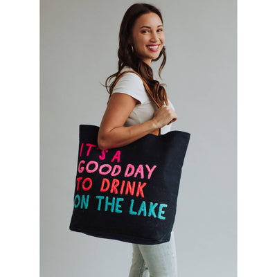 A smiling woman with long brown hair holds the Panache Apparel Co. Navy Drink On The Lake Tote, featuring vibrant ITS A GOOD DAY TO DRINK ON THE LAKE text. She wears a white cotton shirt and light jeans, set against a plain background.
