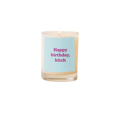 The Birthday Bitch Candle by Talking Out of Turn features a lit white soy-based candle in a glass jar with a blue label that says Happy birthday, bitch in bold pink letters. Made with cotton wicks for a clean, paraben-free burn.