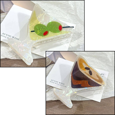 Two lightweight, acrylic Martini Hair Claw Clips by SoapElenGlen—one with green olives and one with a brown liquid design—are shown on a textured white surface with a note underneath. Each clip measures 7CM across.