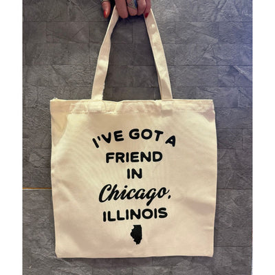 A hand holds the Mugsby "I've Got a Friend in Chicago" Tote, a 15x16 bag made from 10 oz cotton, with black text and an Illinois state silhouette. The background is a gray textured surface.