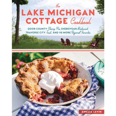 The Lake Michigan Cottage Cookbook by Hachette Book Group features a cherry pie with whipped cream on the cover, set on a picnic table with Adirondack chairs and a lake view—showcasing recipes and cottage life.