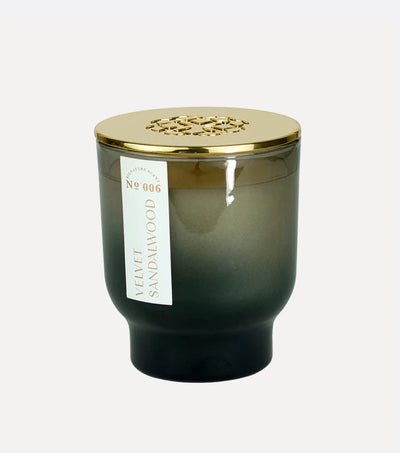 The Velvet Sandalwood 7 oz Maeve Glass Candle by Elum, in a gradient dark green glass with a gold lid and cut-out pattern, exudes serenity. Its label reads Velvet Sandalwood No 006, inviting relaxation against a white background.