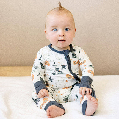 A blue-eyed baby with a blonde mohawk sits on a white bed, wearing the Little Joy Co. Boo Crew Bamboo Zippy Romper, featuring Halloween prints like pumpkins and black cats, plus a YKK zipper for added charm.