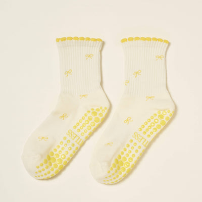 The Mini Bow Petal Grip Sock [Butter] by Effortless Club features white organic cotton, yellow scalloped cuffs, small yellow bow designs, and yellow non-slip grips on the soles, shown on a light background.