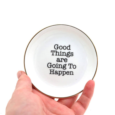 A hand holds the Lenny Mud "Good Things are Going to Happen" Ring Dish, featuring bold black text in the center and a 22K gold rim—an encouraging porcelain gift for jewelry.