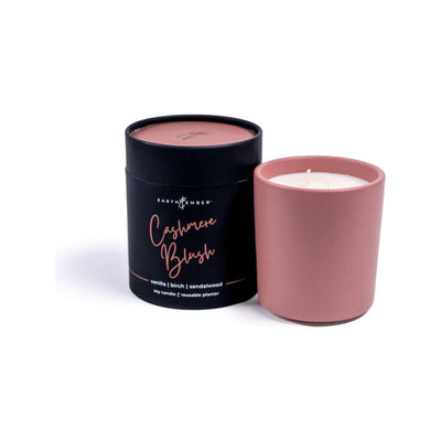 The Earth & Ember 13 oz Candle: Cashmere Blush by Milkhouse Candle Company features a pink double-wick candle with premium fragrance oils and notes of vanilla, birch, and sandalwood in a matching container beside its black cylindrical box.