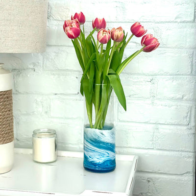 The Artful Nests Glass Flower Vase with Resin Ocean Waves, holding pink tulips and green leaves, sits on a white table next to a candle and lamp against a painted white brick wall.