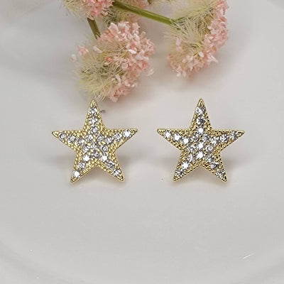 BETTYOH INC Star Post Earrings: gold-tone posts encrusted with sparkling cubic zirconia, shown on a white surface with soft pink flowers in the background.