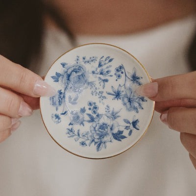 A person with manicured nails holds a Sweet Water Decor Blue Delft Jewelry Dish, a small round ceramic dish adorned with intricate blue Delft rose and flower designs and a gold rim.