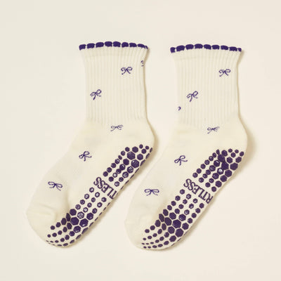 The Effortless Club Mini Bow Petal Grip Sock [Indigo] features organic cotton, purple bow patterns, scalloped edges, cushioned footbed, purple non-slip dots, and “PRINCESS” printed in caps.