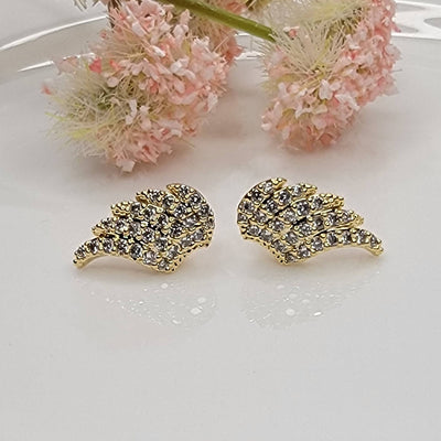 The Wing Post Earring by BETTYOH INC, featuring gold wing shapes embellished with sparkling cubic zirconia, is displayed on a white surface with soft pink flowers in the background.