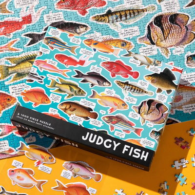 The Judgy Fish 1000 Piece Jigsaw Puzzle by Chronicle Books sits atop a half-finished fish puzzle, its box displaying illustrated fish with funny captions. Loose pieces and another fish-themed surface are nearby.
