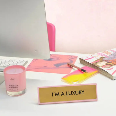 A desk features a computer monitor, stack of magazines, pink pen and phone case, a Flamingo Candles Black Tea & Almond candle, and an Im a Luxury desk plate. The decor includes pink accents in a stylish design ideal for motivational gifts.