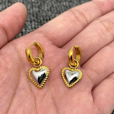 A hand holds two Mio Queena Two Tone Heart 18K Gold Plated Huggie Earrings, each with a non-tarnish silver heart and gold beaded border, against a textured gray background.