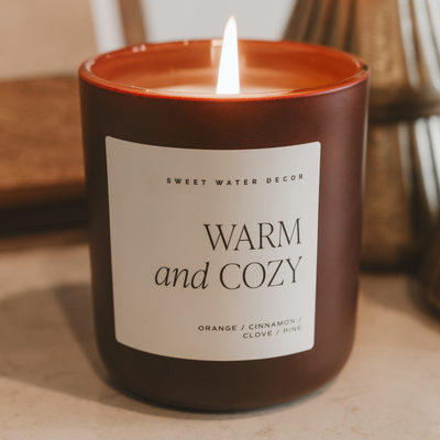 The Sweet Water Decor Warm and Cozy 15 oz Soy Candle in a matte jar sits on a light surface, filling the air with orange, cinnamon, clove, and pine notes for a festive, inviting Christmas scent.