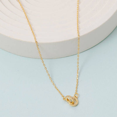 The Gold Dipped Linked Mini Cz Ring Pendant Necklace by Fame Accessories features a delicate design with a simple clasp, shown on a light blue surface next to a white, round, textured object.