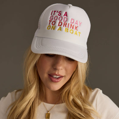 A woman with long blonde hair wears the Panache Apparel Co. White Drink On A Boat Trucker Hat, featuring the vibrant embroidery Its a good day to drink on a boat. Wearing a white top, she looks slightly downward and smiles effortlessly.