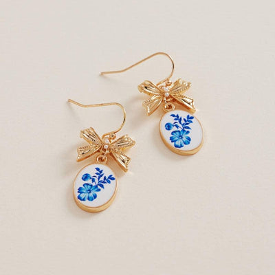 Designature New Yorks Oval Pattern Enamel Charm Metal Bow Hook Earrings feature gold, bow-topped dangles with oval white pendants and blue floral designs, adding elegant charm to any outfit. Displayed on a light beige background.