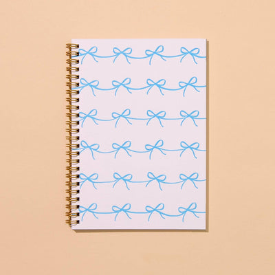 The Bows Letterpress Spiral Notebook by And Here We Are features a cover with rows of light blue bows on a white background, set elegantly against a pale beige surface.