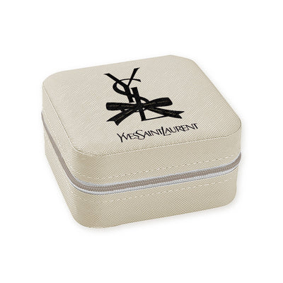 The Travel Jewelry Case - YSL Bow by Toss Designs is a compact, square, vegan leather box in a light shade featuring the iconic black Yves Saint Laurent logo on the lid with a zipper and thoughtfully designed compartments ideal for travel.