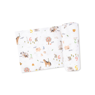 The Angel Dear Farm Sounds Bamboo Knit Swaddle Blanket features colorful farm animal illustrations, numbers, and fun sounds like "moo" and "oink." Shown partially folded with a rolled-up section on the right.