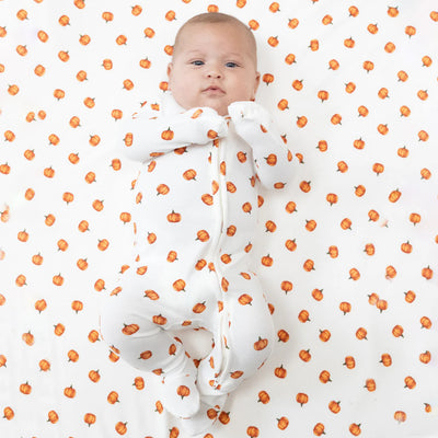 A baby with light skin looks up at the camera, lying on a white blanket with orange pumpkins and wearing the Angel Dear Little Pumpkins Ribbed Two-Way Zipper Footie, featuring a matching pumpkin print.