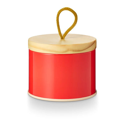 The Aperol Crush Sun Tin Candle by Illume, a round red tin with a light wooden lid and yellow loop handle, evokes the zest of an Aperol Spritz. Its shiny surface reflects like prosecco bubbles, set against a white background.