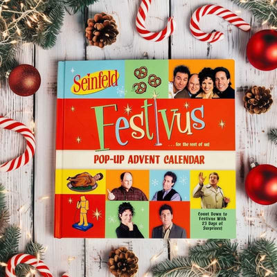 The Seinfeld Festivus Pop-up Advent Calendar by Simon & Schuster rests on a wooden surface with pine branches, candy canes, red ornaments, and string lights—a festive must-have for any Seinfeld fan. UNAVAILABLE FOR SHIPPING.