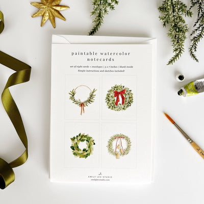 The Wreaths paintable notecards by emily lex studio, featuring four holiday wreath designs, are shown with a green ribbon, watercolor paint tube, paintbrush, and greenery sprigs on a white background.