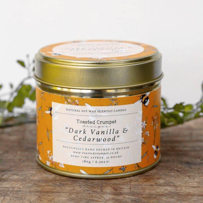 The “Dark Vanilla & Cedarwood Candle” by Toasted Crumpet, with an orange floral tin, gold lid, and white label, rests on wood beside green foliage—a perfect British countryside-inspired gift for candle lovers.