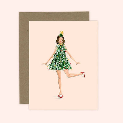 The Jingle Dress Christmas Greeting Card by Almeida Illustrations features a woman in a Christmas tree dress with ornaments and a star headband, standing on one leg and smiling. It comes with a brown envelope.