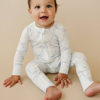 A smiling baby sits on a light tan background wearing the Blue Belle Ribbed Bamboo Zippy Romper by Little Joy Co., a white, long-sleeved bamboo viscose outfit with light blue bow patterns, facing forward with relaxed hands and feet.