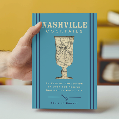 A hand holds Nashville Cocktails by HarperCollins Focus, a blue-covered book with a cocktail glass silhouette. Explore over 100 Music City–inspired drink recipes and discover how to sip like a true Nashville local.