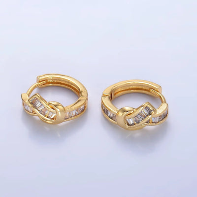 Golden Stella’s Baguette Knot Huggie Hoops are small gold brass hoop earrings, 1/2 inch in diameter, featuring a knot design and rectangular clear stones on the front, shown on a plain light surface.