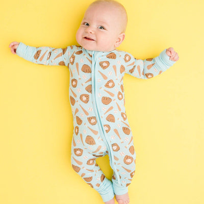 A smiling baby lies on a bright yellow background, wearing the Little One Shop Home Run Blue Bamboo Sleeper with playful lion print, matching cuffs, and fold-over mittens for extra comfort.