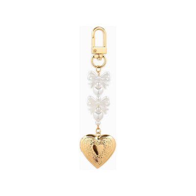 The Sessori Bow Pearl Heart Locket Bag Charm features a gold-tone heart locket, two white pearl-like beads, and two clear bow-shaped charms, all hanging from a gold clasp.