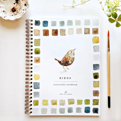 Emily Lex Studios Birds Watercolor Workbook showcases beginner-friendly illustrations with a captivating bird on the cover amidst colorful paint swatches. A paintbrush, small ceramic dish, and green leaves complement this inviting watercolor paper haven.