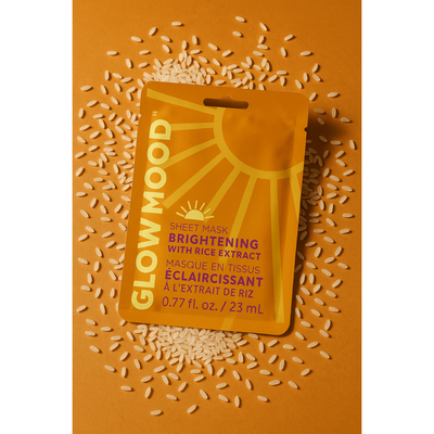 A vibrant orange package of Glow Mood’s Brightening Sheet Mask with Rice Extract sits on a matching surface amid rice grains. The vegan, cruelty-free mask features “Brightening with Rice Extract” in English and French.