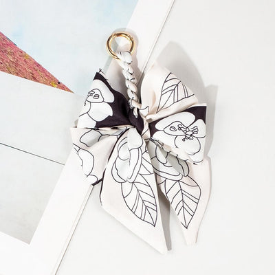The Judson & Company Scarf Bow Key Ring features a 6-inch braided fabric loop, gold ring, and a large black-and-white floral bow, shown on a white surface beside a photo or print.