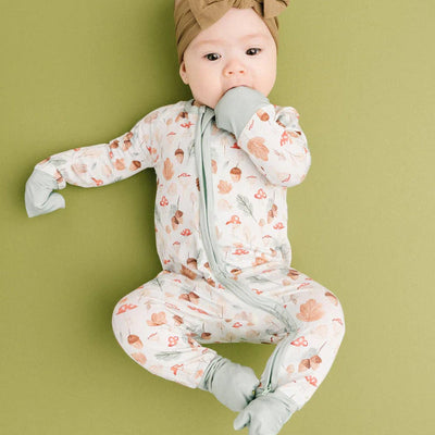 A baby in the Little One Shop Fall Woodland Bamboo Sleeper with fold-over mittens and footies lies on a green surface, wearing a brown bow headband and sucking on their hand.