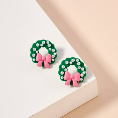The Frem Pearl Wreath with Pink Bow Earrings feature green enamel wreaths adorned with white beads and pink bows, perfect for festive flair. Shown on a white and peach surface.