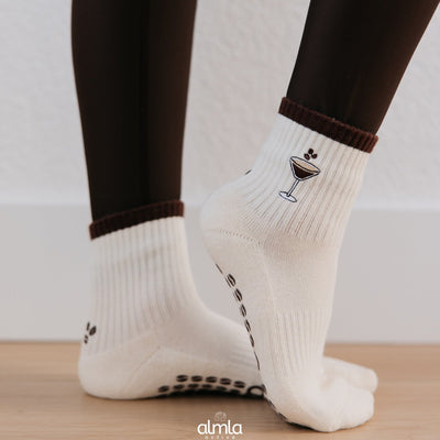 A person stands on a wooden floor wearing Almla Active's Espresso Martini Pilates Socks—white ribbed socks with brown cuffs, a cocktail glass design, and non-slip grips on the soles.