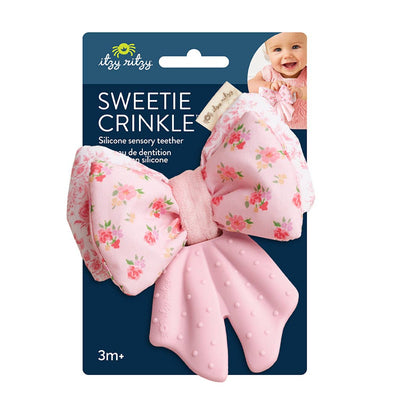 The Itzy Ritzy Bow Sweetie Crinkle™ Toy is a pink floral bow-shaped silicone teether with textured ends, ideal as a sensory toy for babies 3 months+. Packaging features a smiling baby in the top right corner.