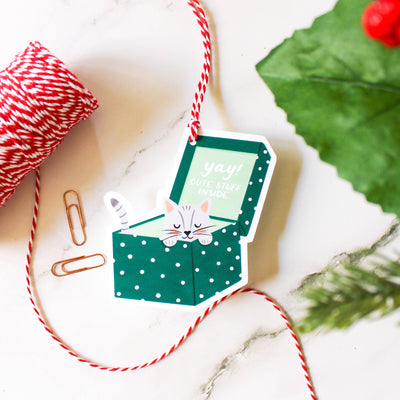 Decorative gift tag with a cat illustration on a marble surface with string and paper clips.