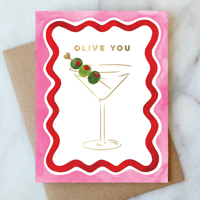 The Olive You Martini Card by Abigail Jayne Design sits on a marble surface, featuring a watercolor martini glass with three olives. Its framed by pink and red waves, with Olive You elegantly shimmering in gold digital print above the glass.