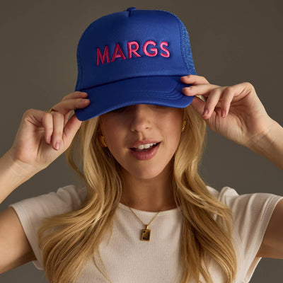 A woman adjusts her Blue Margs Trucker Hat by Panache Apparel Co., featuring MARGS in pink lettering. Her long blonde hair flows gracefully, enhanced by a white top and gold necklace. The hat offers breathable mesh and an adjustable snapback for ultimate comfort.