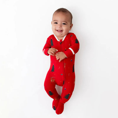 A smiling baby in the Bamboo Little Reindeer Zippered Footie, featuring Christmas tree and reindeer patterns, lies on a white background.