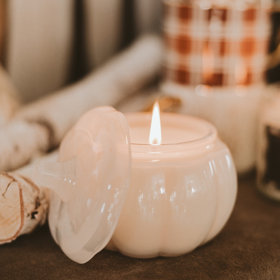 The Warm and Cozy Soy Candle - 20oz Pumpkin Jar by Sweet Water Decor sits lit on a table, its white pumpkin shape and cozy fragrance creating an inviting autumn vibe amid soft-focus fall decor.