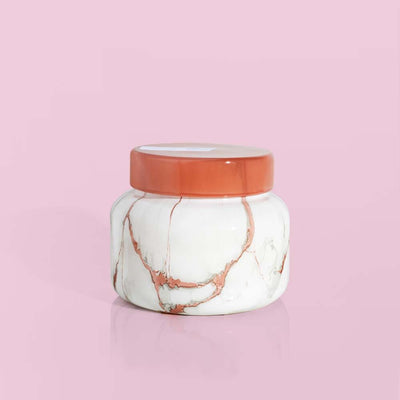 A small Capri Blue Havana Vanilla Modern Marble Signature Jar Candle, with a sleek white and pink marble design and a pink lid, sits elegantly on a solid pink background, infusing the air with serenity.